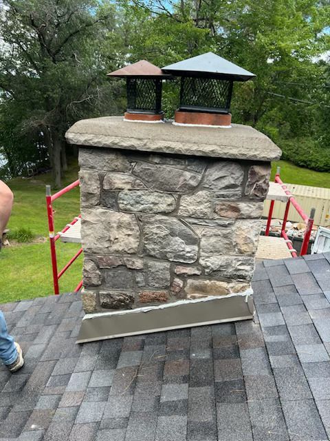 Stone chimney on a shingled roof, with two metal chimney caps and roof scaffolding in the background.