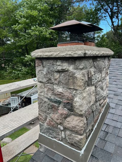 Stone chimney with a metal cap on a shingled roof, framed by trees and a wooden deck railing
