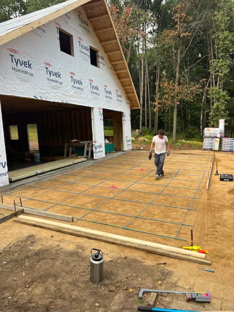 House under construction with rebar grid foundation and a worker on a wooden deck beside a wooded area