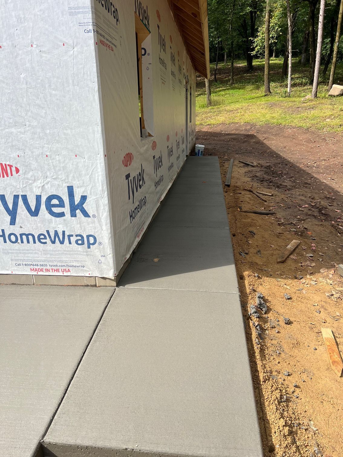New concrete walkway beside a house wrapped in Tyvek, with dirt and woods alongside.