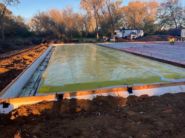 Concrete slab under construction, covered with yellow plastic sheeting beside a dirt lot and trees.