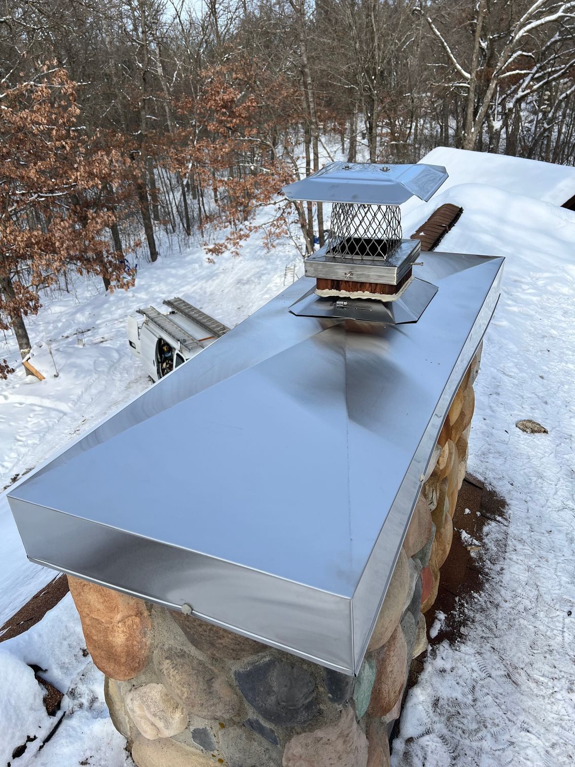 Snow-covered outdoor stone chimney with a shiny metal cap on a winter hillside