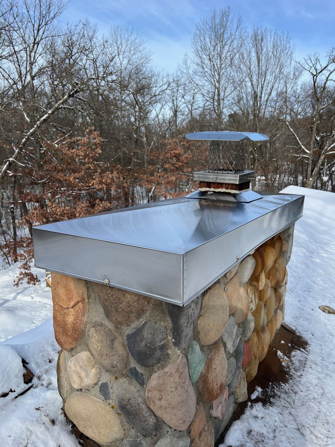 Stone outdoor fireplace with a shiny metal top in a snowy wooded setting