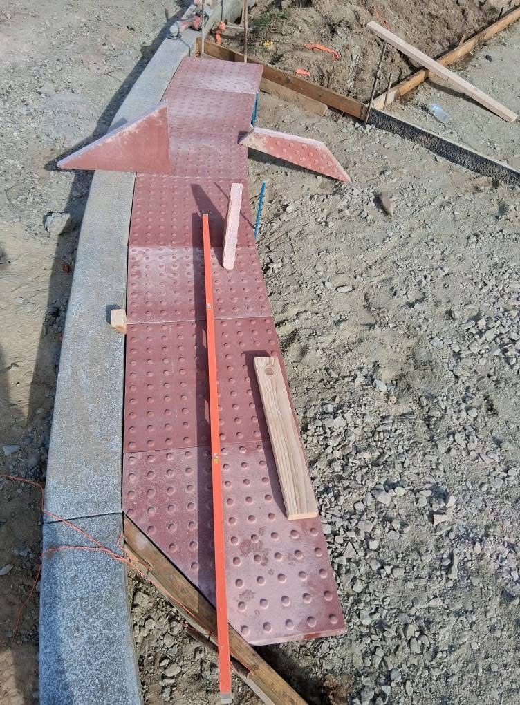 Red metal construction panel with perforations and wooden inserts lying on dirt and gravel.