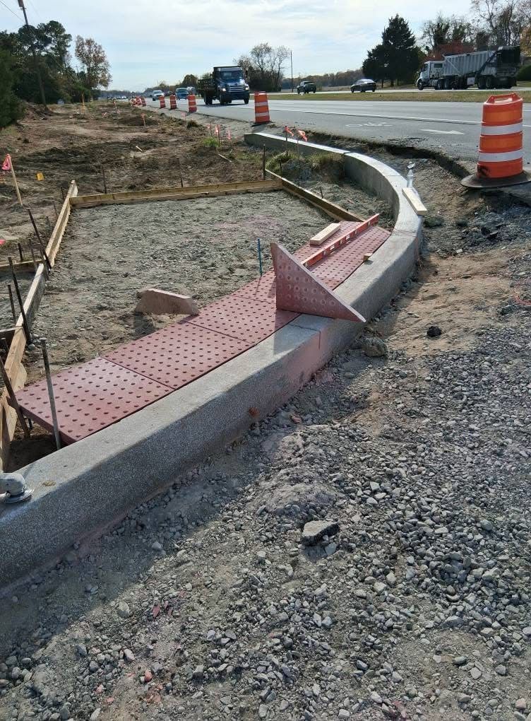 Damaged curb ramp with pink tactile paving at a roadside construction site, bordered by orange barrels