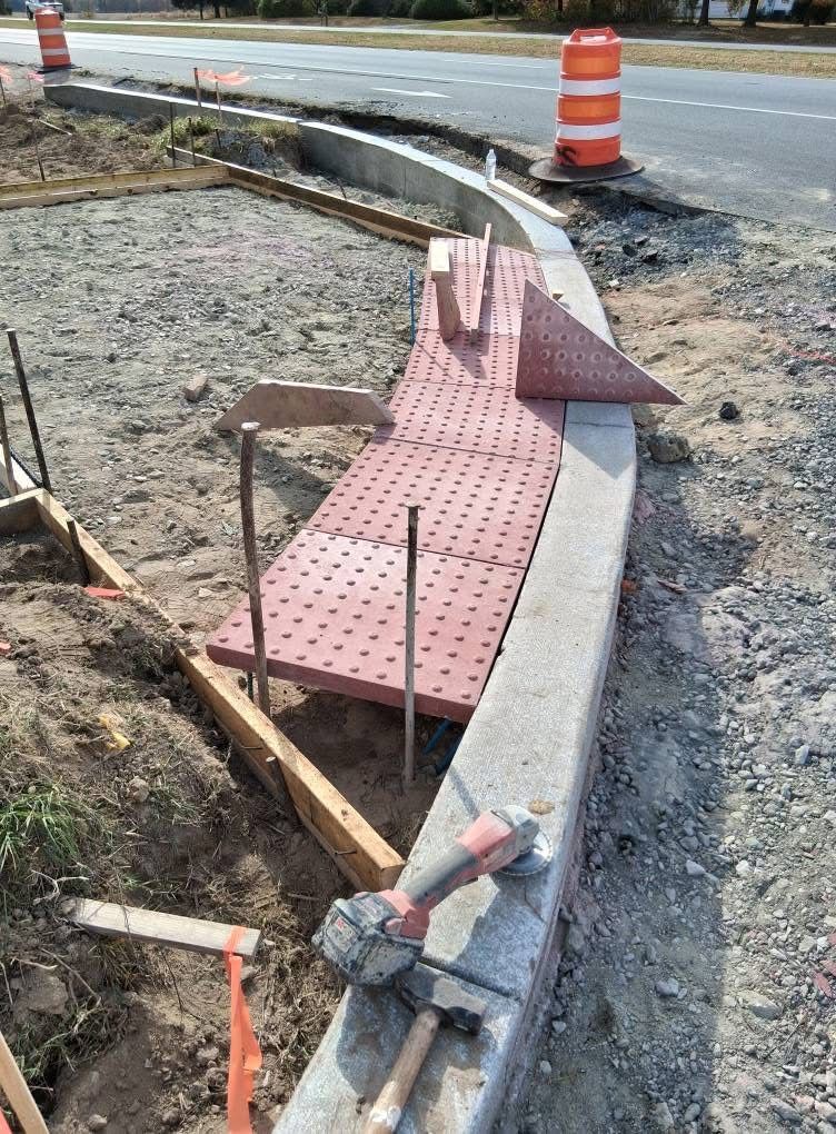 Construction work on a curb ramp with pink tactile paving beside a road and orange traffic barrels