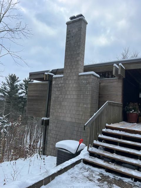 Snowy exterior of a house with a tall chimney and wooden stairs leading to a deck