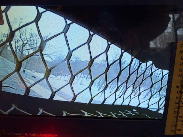 View through a mesh fence toward snowy mountains under a blue sky, with a dark roof edge overhead.