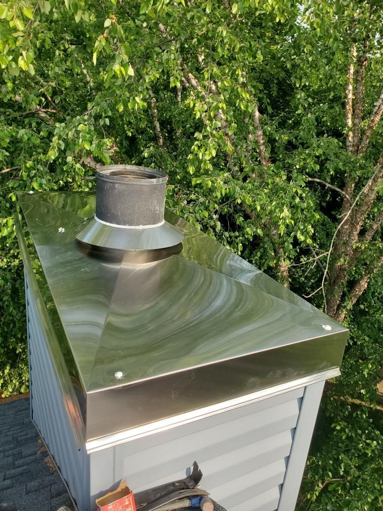 Metal chimney cap on a silver rooftop against green trees.