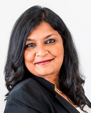 Woman with dark hair smiling, wearing a black blazer and a pearl necklace, against a white background.