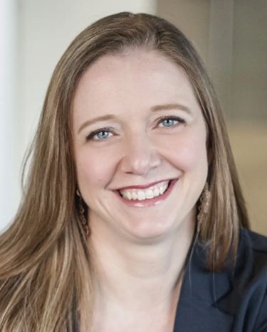 Smiling woman with light brown hair in a dark blazer against a neutral background