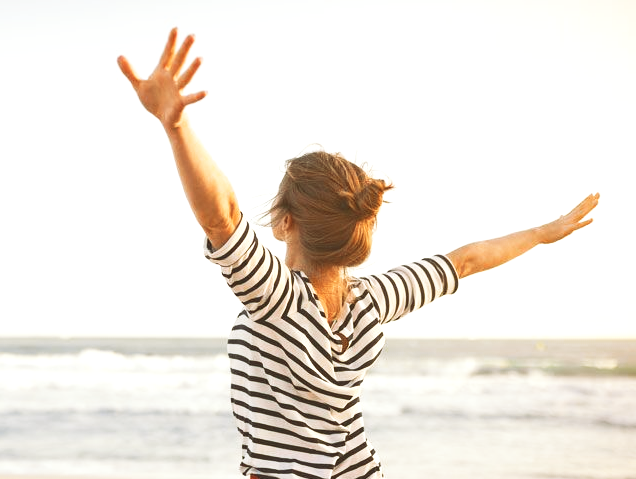 Person in a striped shirt with arms outstretched, facing the ocean on a beach at sunset.