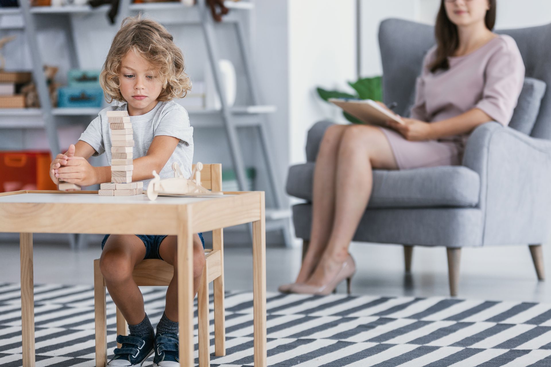 Child writing at a wooden table while an adult sits blurred in a living room background