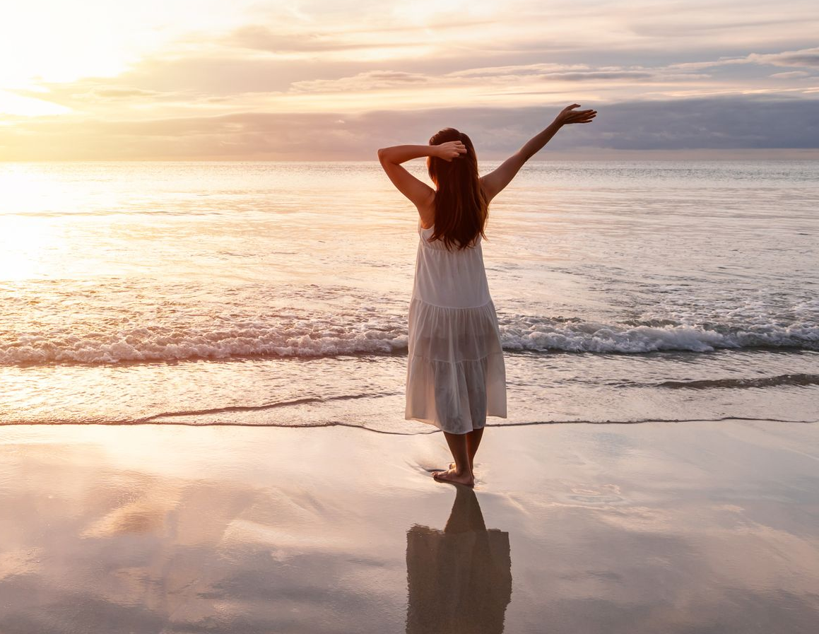 Person in a white dress standing on a beach at sunrise with arms raised toward the sea