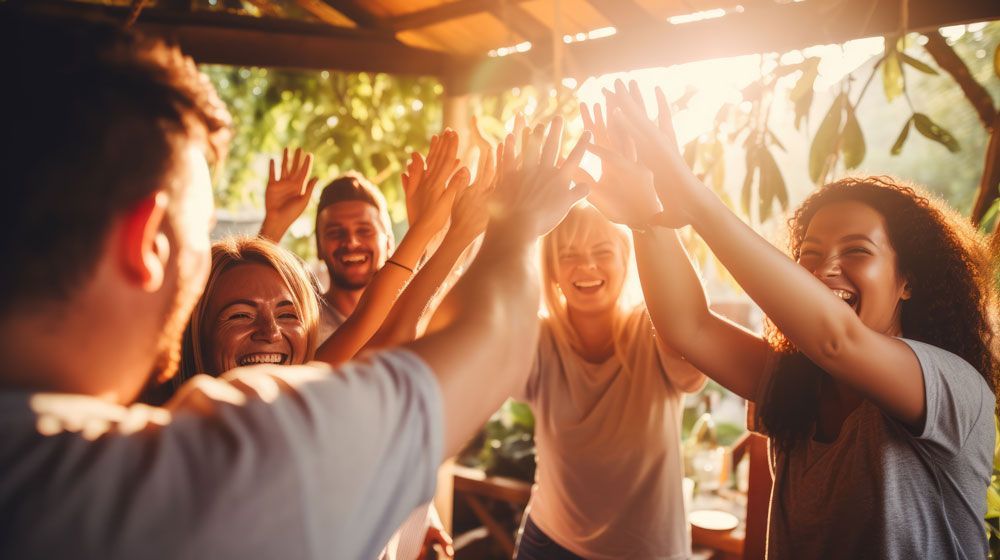 Group of friends high-fiving outdoors under warm sunset light, smiling around a table