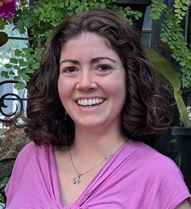 Smiling woman in a pink top standing outdoors with greenery and a wrought-iron gate behind her