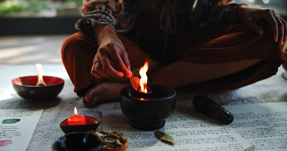 Person lighting a small oil lamp beside bowls and ritual items on a paper-covered floor