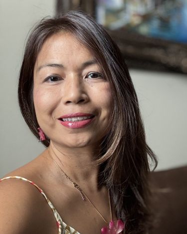 Smiling woman with long dark hair in a pink top, seated indoors with a softly blurred background