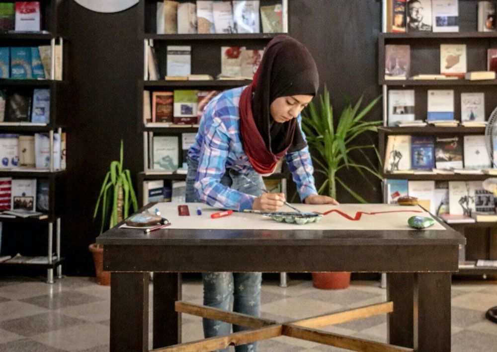 Person leaning over a worktable in a craft or science room, arranging materials with shelves behind.