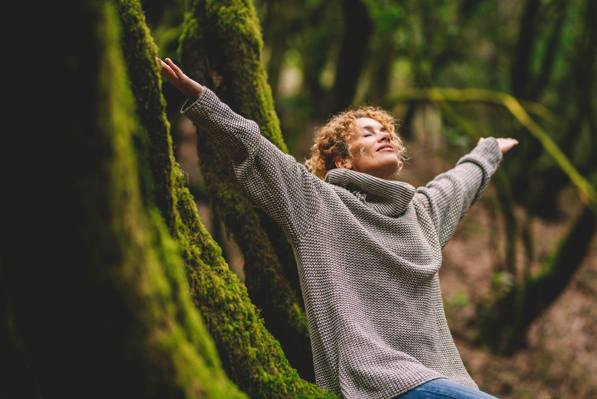 Person in a gray sweater with arms raised in a lush green forest.