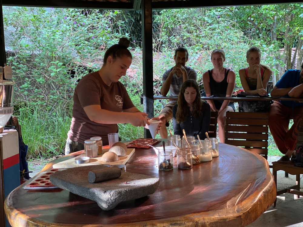 Person preparing food at a round wooden table while others watch in an open-air pavilion.
