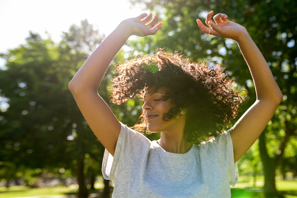 Person dancing outdoors in sunlight with arms raised, curly hair, and a white shirt