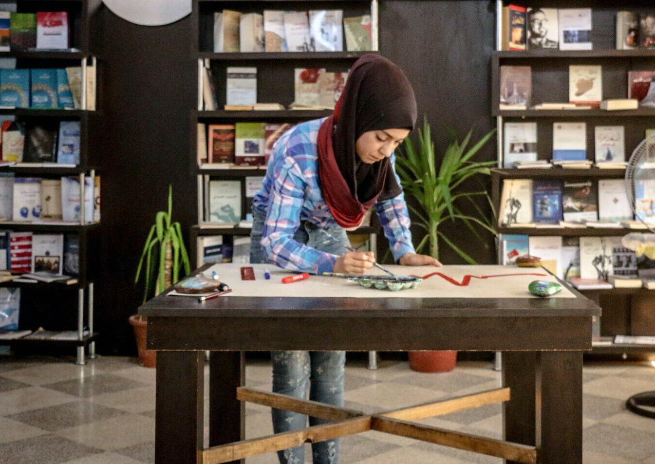 A woman in a hijab is sitting at a table in front of a bookshelf.