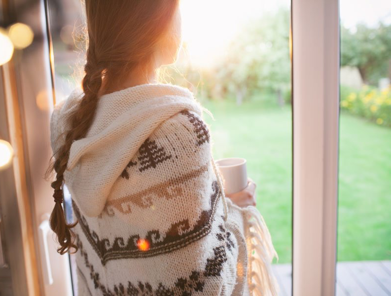 Person in a patterned blanket holding a mug by a sunlit window, looking out onto a green garden.