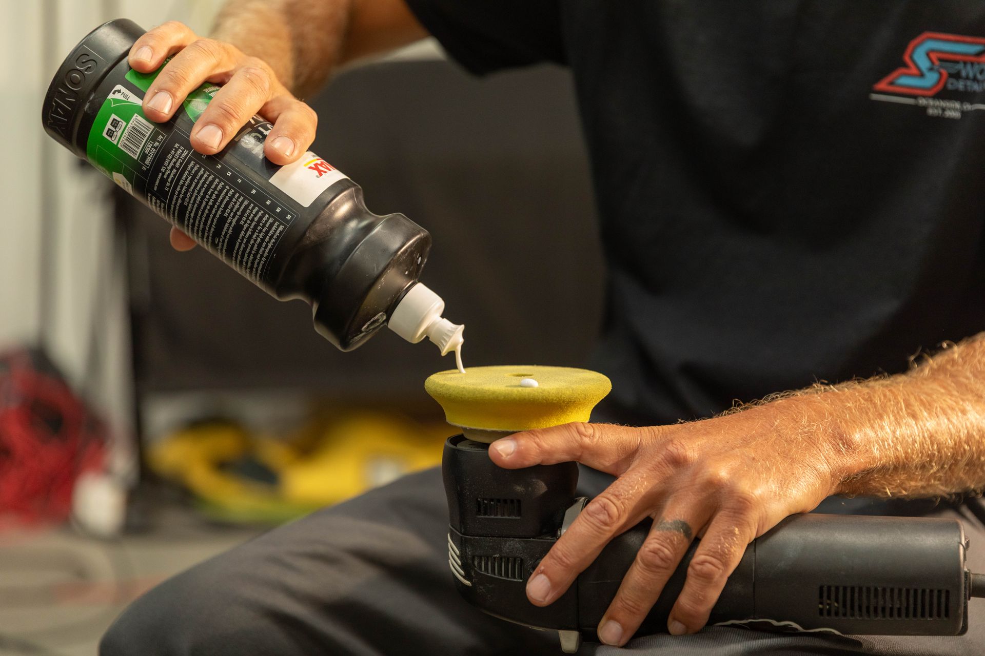 Hands applying polish to a black car with a handheld buffer in a garage