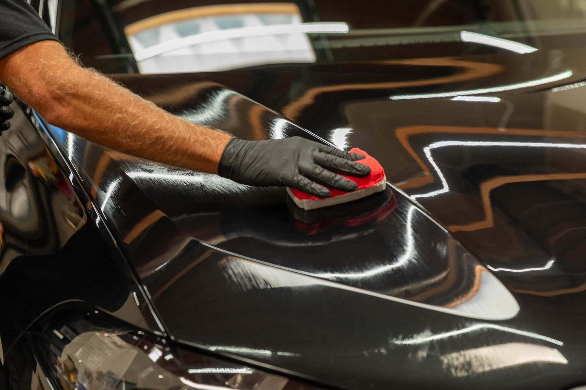Gloved hand polishing a glossy black car hood with a red applicator pad