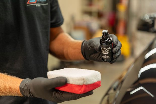 Gloved hands holding a spray bottle and a red-and-white microfiber applicator in a workshop