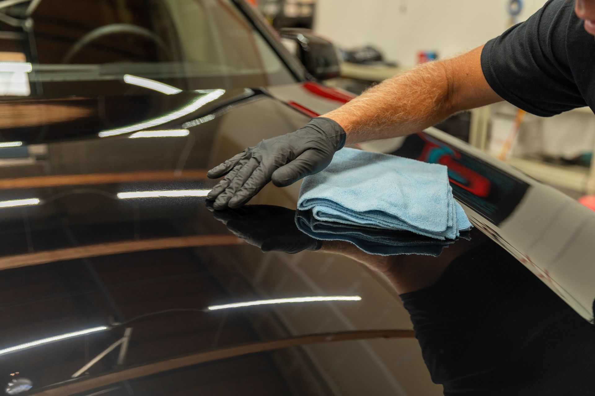 Gloved hand wiping a glossy black car hood with a blue microfiber cloth