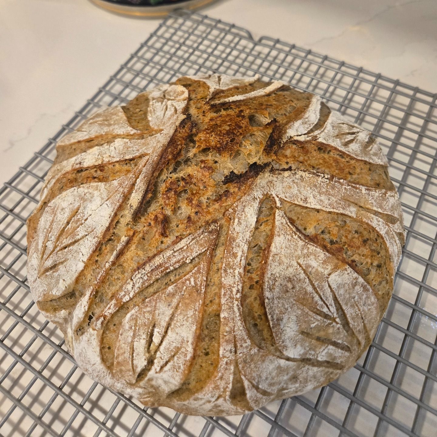 Round artisan loaf of bread with leaf-patterned scoring, cooling on a wire rack.