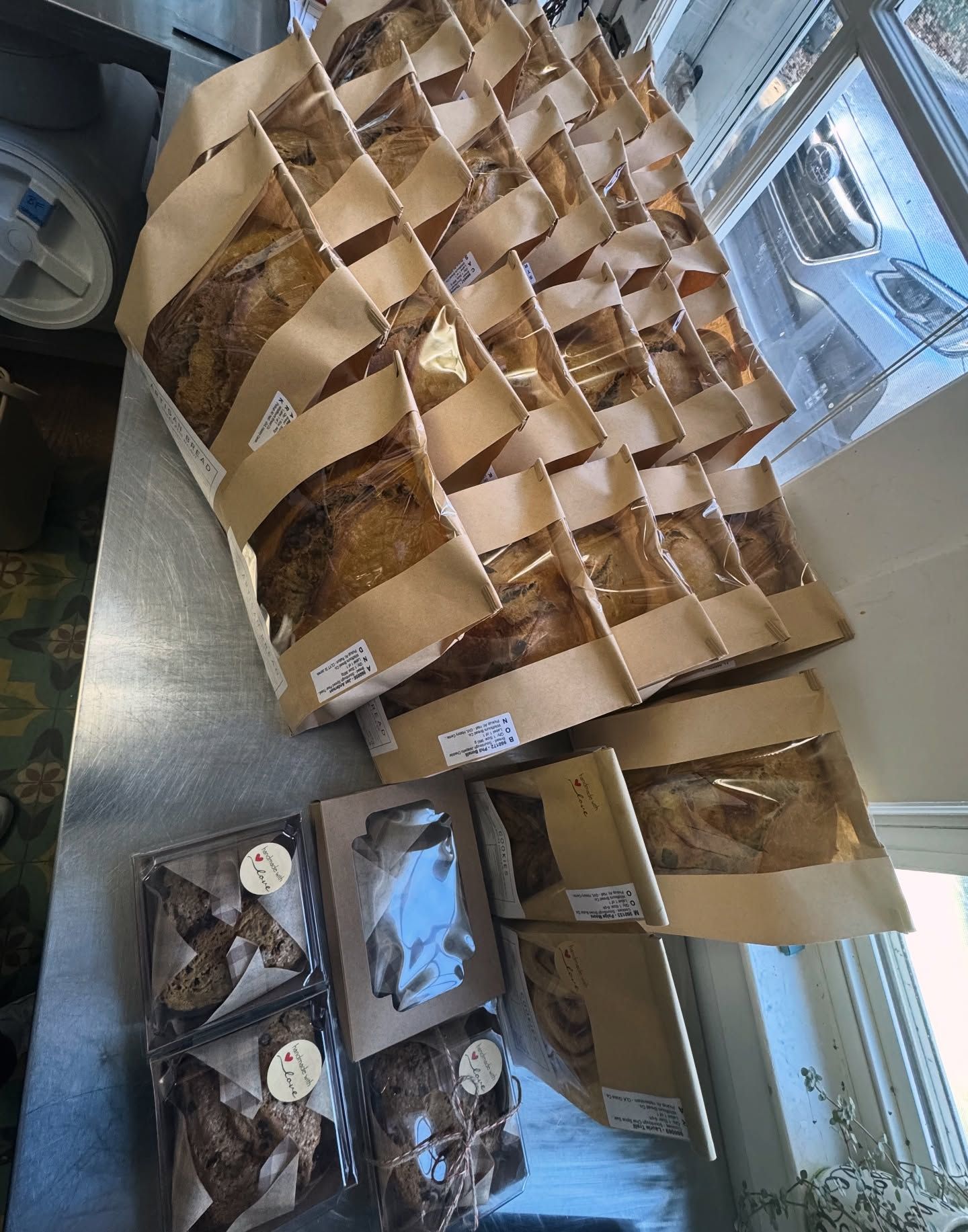 Paper bags of baked goods and packaged cookies displayed on a counter by a window.