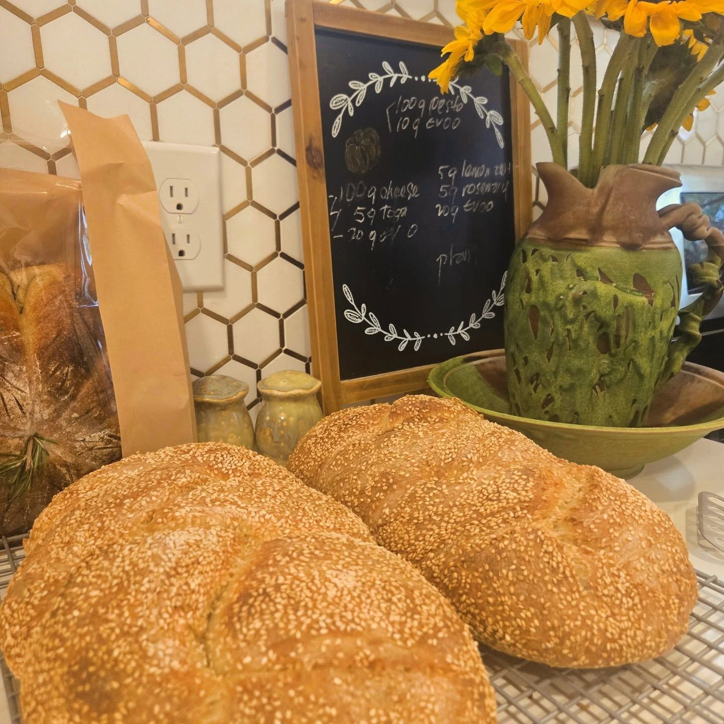 Sesame bread loaves on a counter beside a chalkboard sign, flowers, and a green vase