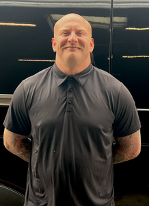 Smiling bald man in a dark polo shirt, standing with hands behind his back against a black background