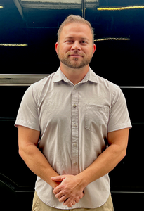 Man in a light gray shirt standing with hands clasped against a dark background