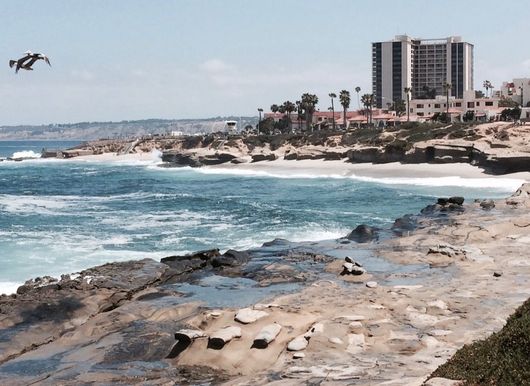 Rocky shoreline with crashing waves and a seaside resort building under a hazy sky