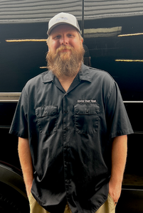 Man standing in a black shirt and cap in front of a dark vehicle, with a long beard.