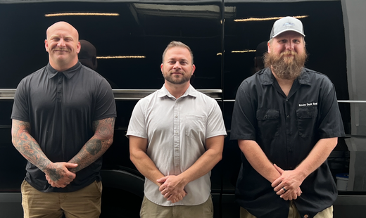 Three men standing side by side in front of a black vehicle, facing the camera.