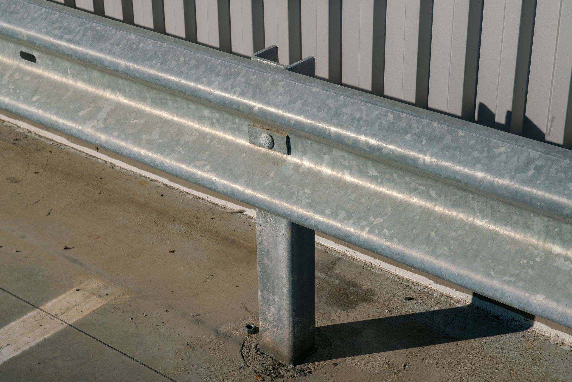 Metal guardrail along a concrete surface, supported by a vertical post; a gray fence is in the background.