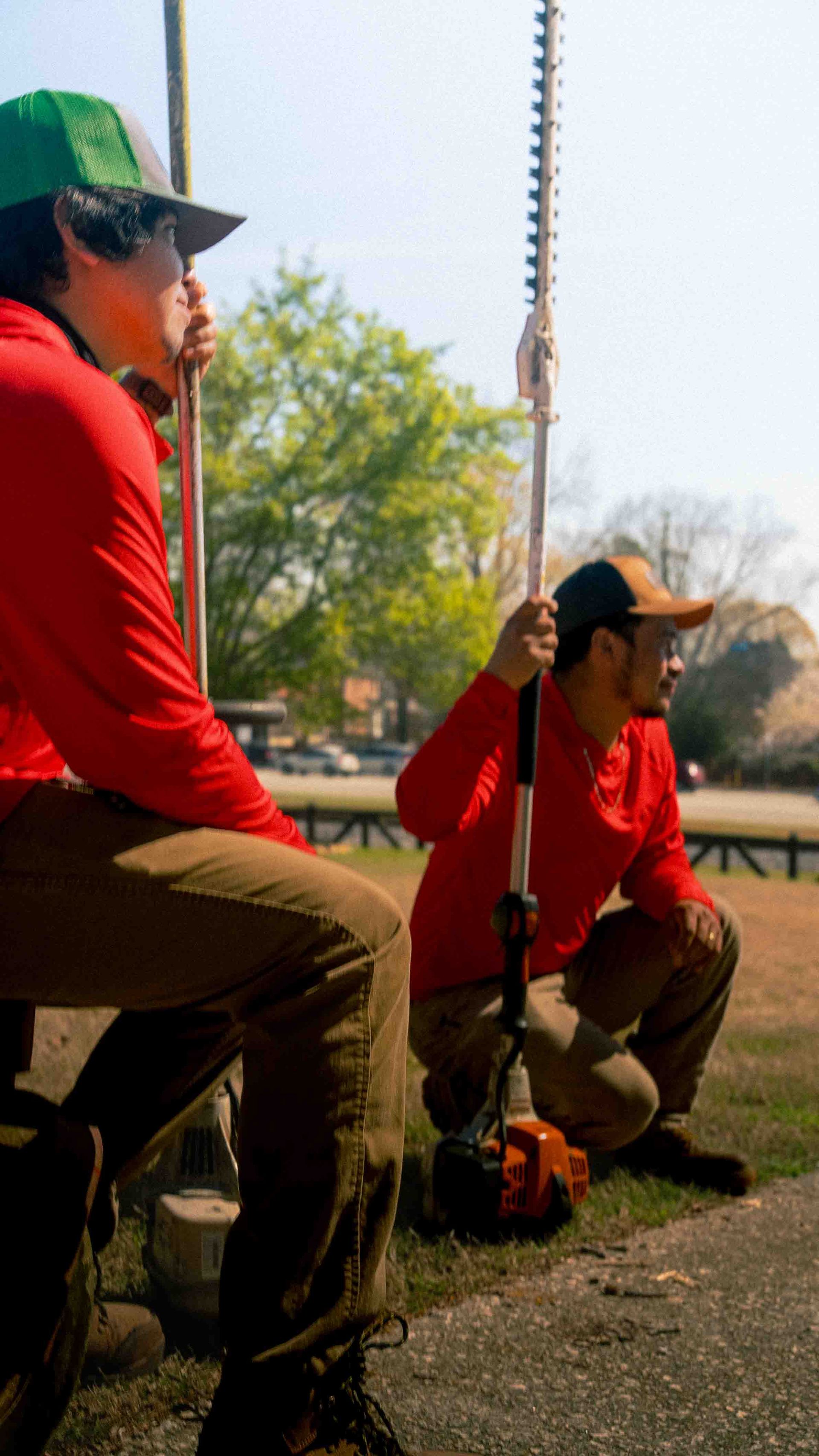 Two workers in red shirts and caps crouch outdoors, holding long-reach hedge trimmers in a park setting.