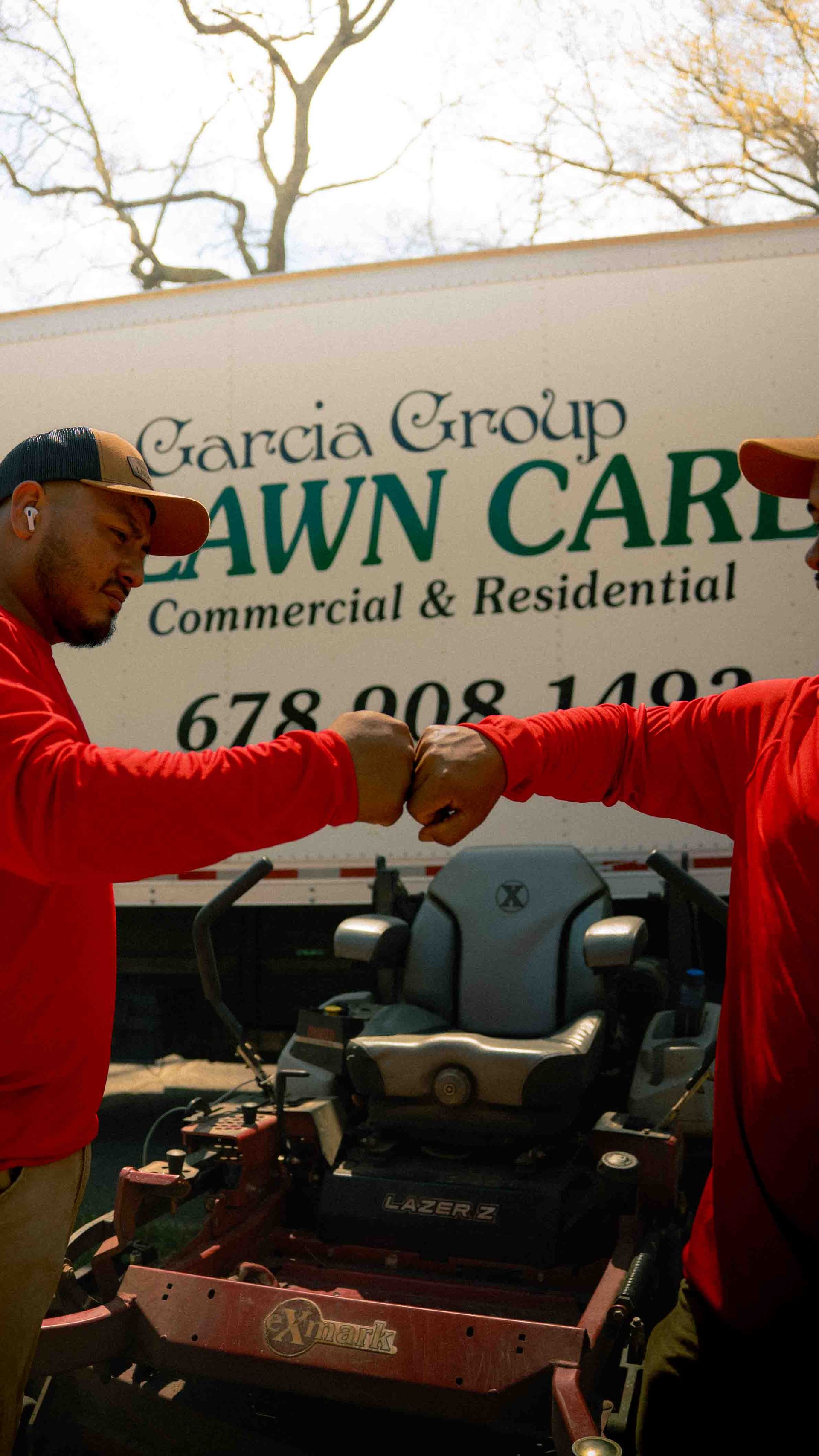 Two workers in red long-sleeve shirts fist bump in front of a Garcia Group Lawn Care truck and a zero-turn mower.
