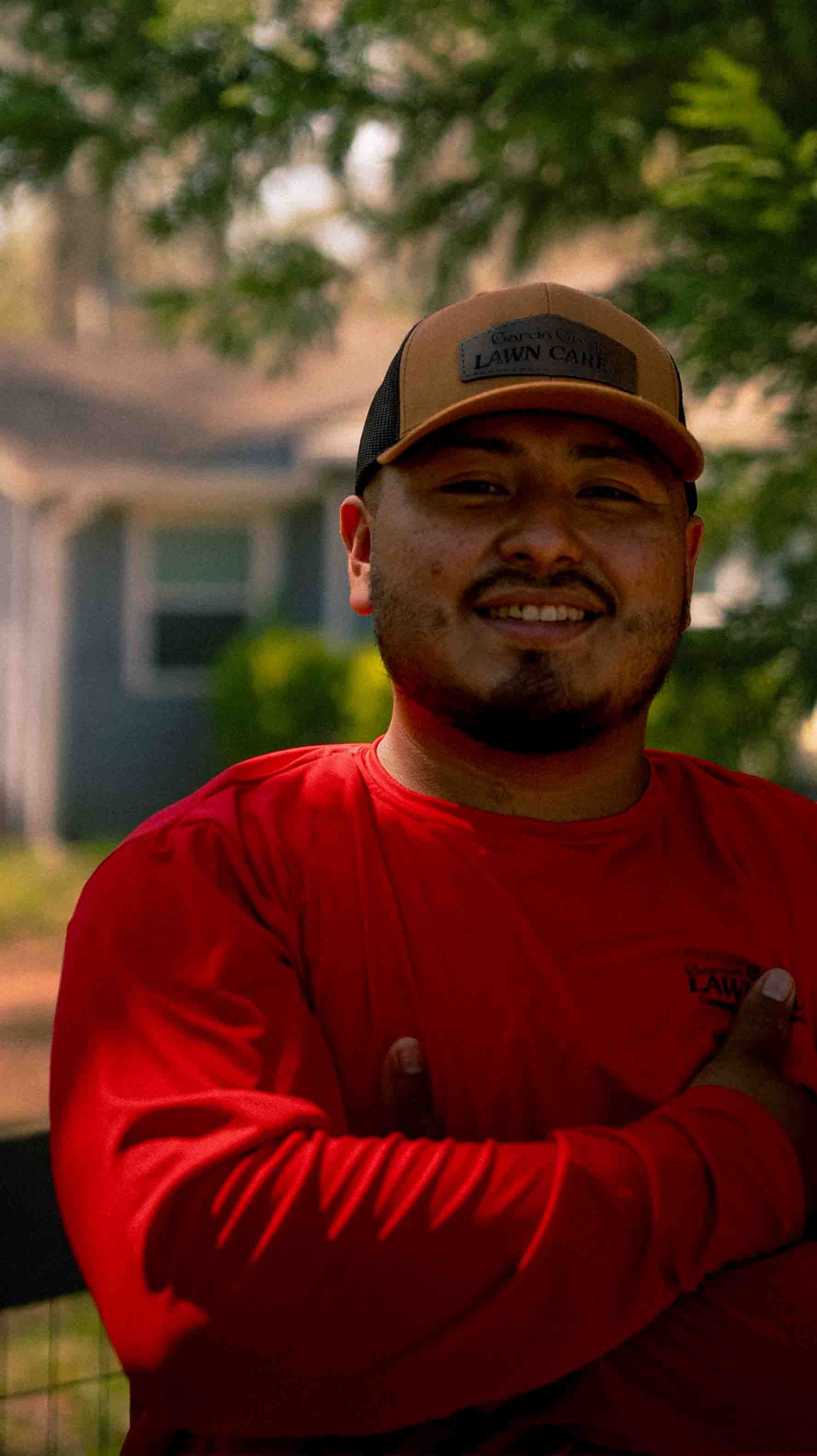 A person wearing a red long-sleeve shirt and a brown baseball cap, standing outside with arms crossed and smiling.