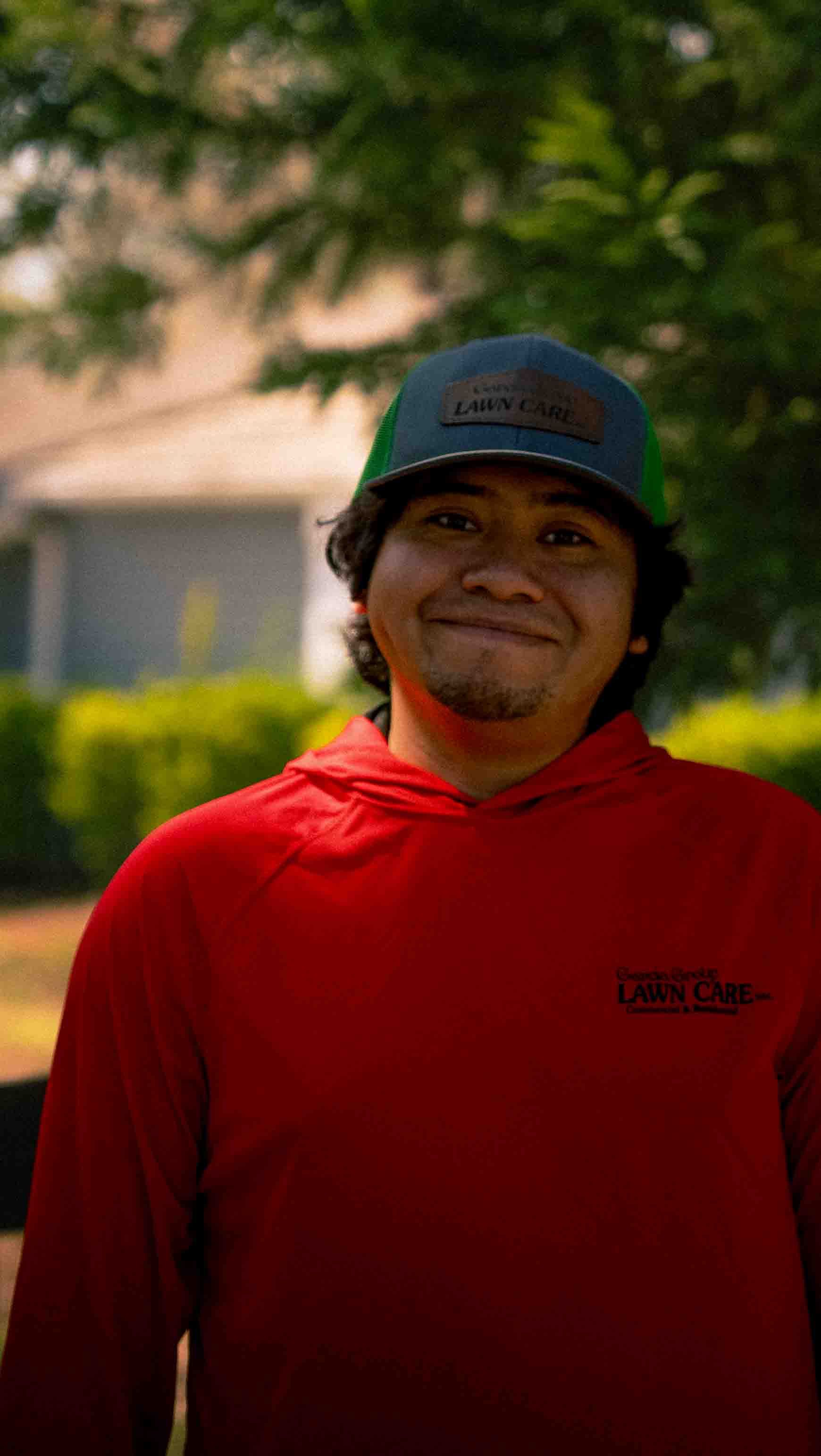 A smiling person wearing a red hooded sweatshirt and a green baseball cap, standing outdoors in front of greenery.