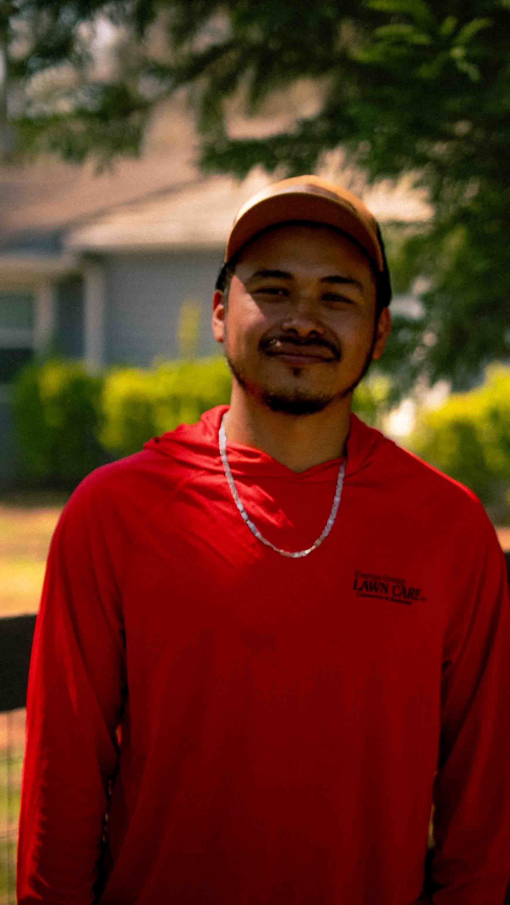 A person wearing a tan baseball cap and long-sleeved red shirt with a chain necklace, standing outdoors in front of trees.