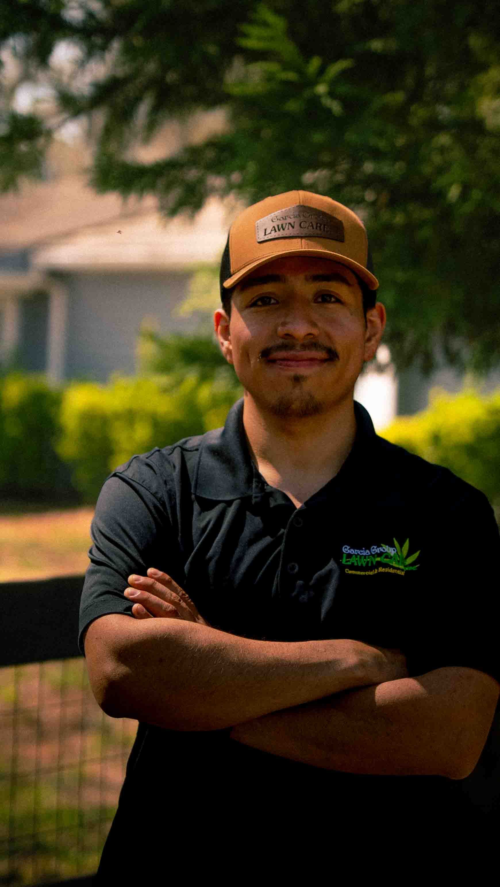 A person with a mustache and a tan cap crosses their arms, wearing a black polo shirt, standing outdoors by a fence.