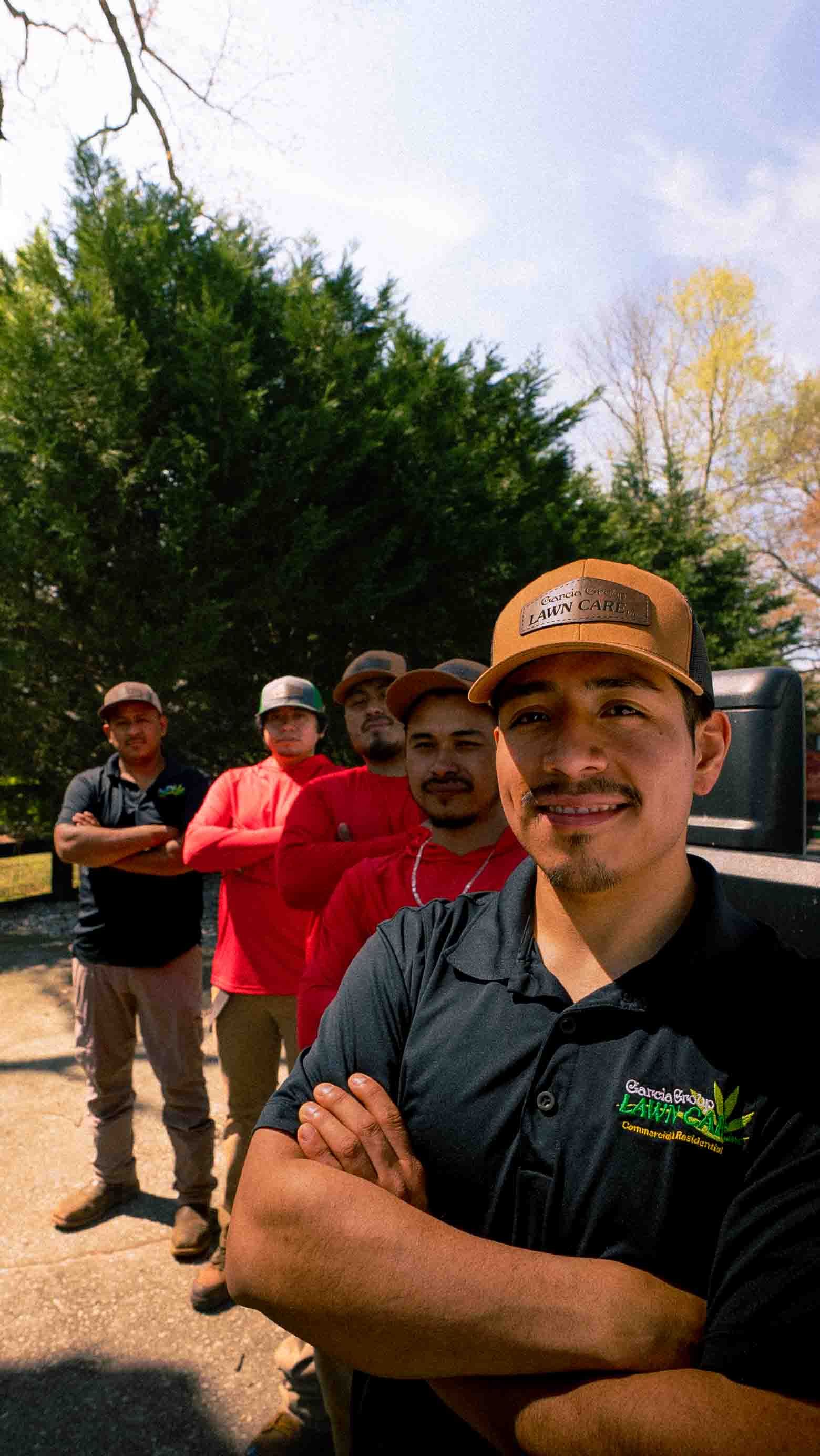 A line of five workers in uniform shirts and caps stand with arms crossed outdoors in front of a tree line.