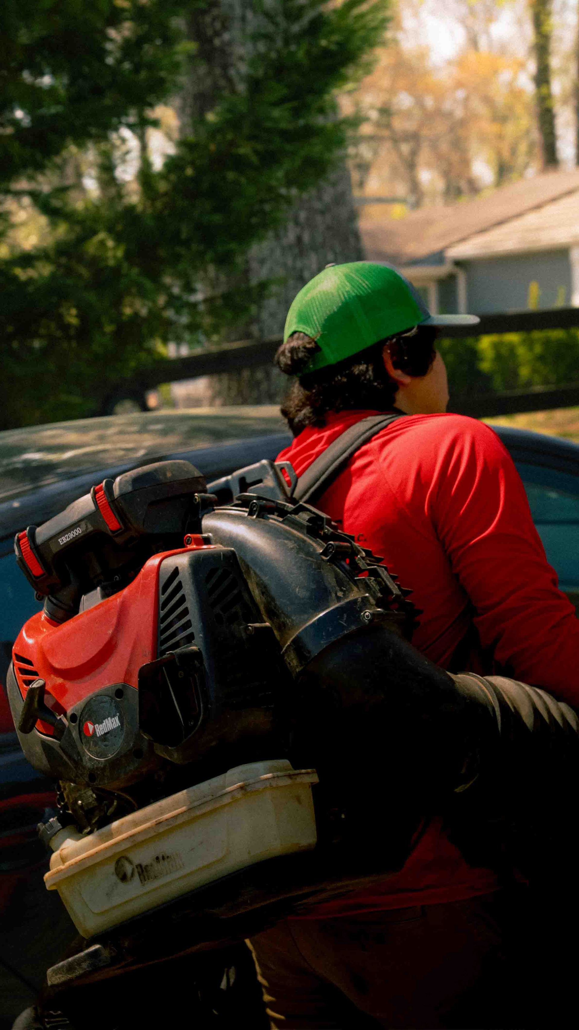A person wearing a bright green cap and red shirt carries a red gas-powered backpack leaf blower outdoors.