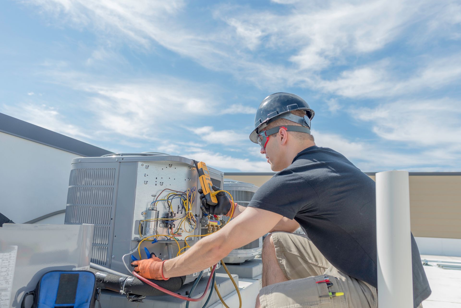 A person in a hard hat repairs an air conditioning unit on a rooftop under a blue sky.